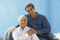 Senior man in chair, adult son behind, clasped hands - Manoj Adhikari