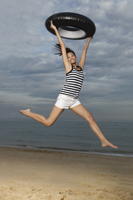 Young woman jumping on beach, holding up inner tube - Yukmin