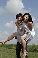 young man carrying a woman on his back laughing with blue sky and clouds in background. - Yukmin