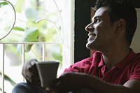 Indian man looking out window smiling holding coffee cup - Alex Mares-Manton