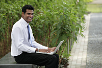 Indian man sitting on park bench working on laptop and smiling. - Alex Mares-Manton