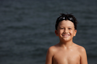 Young boy standing in front of ocean smiling with goggles on his head - Yukmin