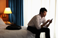 young man sitting on bed in hotel room texting on phone - Yukmin