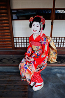 Geisha in red Kimono sitting in front of house - Travelasia