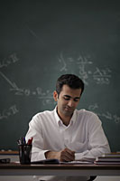 Young man sitting at his desk writing with chalk board in background. - Alex Mares-Manton