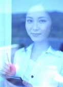 Young woman holding palmtop, standing behind window, smiling - Jade Lee
