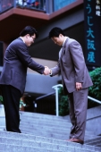 Two male executives bowing while shaking hands on stairway - Keith Brofsky