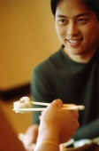 Young man smiling, hand holding chopstick with sushi in foreground - Keith Brofsky