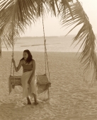 Woman sitting on swing under coconut tree looking off camera, sea in background - Jack Hollingsworth
