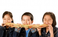 Three young children (two girls, one boy) eating large sandwich, white background. - Jade Lee