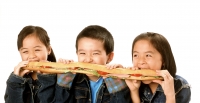 Three young children (two girls, one boy) eating large sandwich, white background. - Jade Lee