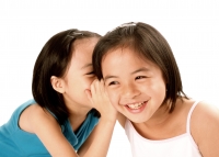Two young girls whispering to each other, laughing, white background. - Jade Lee