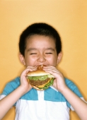 Young boy eating large hamburger, yellow background. - Jade Lee