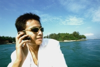 Young man with sunglasses using cellular phone, blue sky and water in background - Eric Ceret