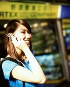 Young woman using cellular phone, shops in background. - Jack Hollingsworth