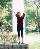 Young woman standing with arms outstretched, fountain in background. - Jack Hollingsworth