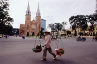 Vietnam, Ho Chi Minh City, Notre Dame Cathedral, woman crossing road with fruit baskets in foreground. - Steve Raymer