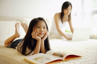 Mother and daughter in bedroom, mother using laptop, daughter lying down with book open in front of her - Alex Microstock02