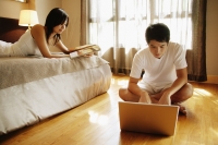 Young man using laptop, young woman lying on bed reading magazine - Alex Microstock02