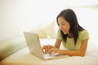 Young woman lying on bed, using laptop, smiling - Alex Microstock02