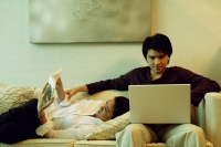 Man sitting on sofa, using laptop, woman lying next to him reading magazine - Alex Microstock02