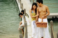 Couple walking on jetty, man carrying picnic basket, people in the background - Jade Lee