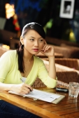 Young woman sitting at table with pen and paper, hand on face - Alex Microstock02