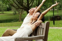 Couple sitting on park bench, looking up, woman with arms outstretched - Alex Microstock02