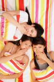 Three young women lying on bed, looking up at camera, hands over mouths - Alex Microstock02