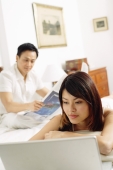 Couple in bedroom, woman lying on bed, looking at laptop, man reading newspaper behind her - Alex Microstock02