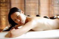 Woman lying on massage table, stones along her back, looking at camera - Alex Mares-Manton