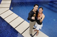 Couple standing by swimming pool, looking up at camera, holding champagne glasses - Alex Mares-Manton