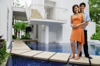 Couple standing outside house, next to swimming pool, looking at camera - Alex Mares-Manton