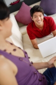 Couple in living room, man lying on sofa with laptop, smiling at woman - Alex Mares-Manton