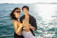 Couple on boat deck, standing next to railing, holding champagne glasses, smiling at camera - Alex Mares-Manton