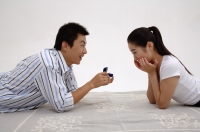 Couple lying on front, face to face, man holding ring box, woman with hands on chin - blueduck