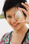 Woman holding shell over one eye, smiling at camera - Alex Microstock02