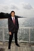 Businessman standing next to sea, hand in pocket, looking at camera - Yukmin