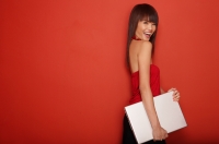 Young woman in red tube top, standing against red wall, carrying laptop - Yukmin