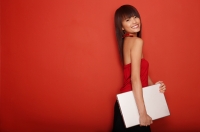 Young woman standing against red wall carrying laptop - Yukmin