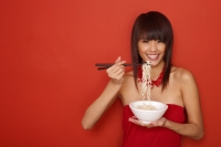 Young woman standing against red wall, holding bowl of noodles, smiling - Yukmin