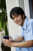 Young man leaning on railing, holding mug, smiling at camera - Yukmin