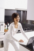 Young woman standing in kitchen, using laptop, smiling at camera - Alex Mares-Manton