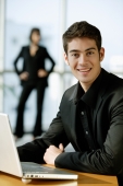 Male executive dressed in black, sitting next to laptop, looking at camera, smiling - Alex Mares-Manton