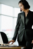 Businesswoman dressed in black, standing, looking at laptop on desk - Alex Mares-Manton
