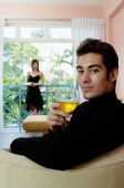 Man sitting in living room, holding a glass of wine, woman in the background - Yukmin