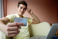 Man at home, using laptop. holding credit card and looking at camera - Yukmin