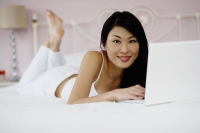 Woman lying on bed, using laptop, smiling at camera - Yukmin