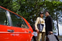 Couple carrying bags, standing next to red car, smiling at camera - Alex Mares-Manton