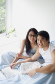 Couple sitting side by side in bedroom with laptop, smiling at camera - Alex Mares-Manton